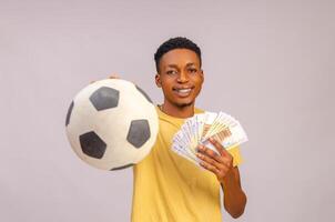 Sport Wette Aufregung, Glücksspiel und Familie Lebensstil Konzept. aufgeregt Afroamerikaner jung Kerl genießen ihr gewinnen, Stehen mit Fußball Ball und viele von Geld foto