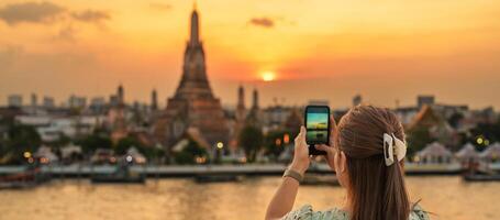 Tourist Frau genießt Aussicht zu wat arun Tempel im Sonnenuntergang, Reisender nehmen Foto zu Tempel von Dämmerung durch Smartphone von Dach Bar. Wahrzeichen und Reise Ziel im Bangkok, Thailand und Süd-Ost Asien