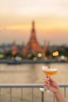 Tourist Hand zeigt an Cocktail Glas während genießen Aussicht zu wat arun Tempel im Sonnenuntergang, Reisender besucht Tempel von Dämmerung in der Nähe von von Dach Bar. Reise Ziel im Bangkok, Thailand und Süd-Ost Asien foto