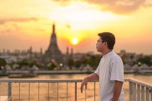 Tourist Mann genießt Aussicht zu wat arun Tempel im Sonnenuntergang, Reisender besucht Tempel von Dämmerung in der Nähe von Chao Phraya Fluss von Dach Bar. Wahrzeichen und Reise Ziel im Bangkok, Thailand und Süd-Ost Asien foto