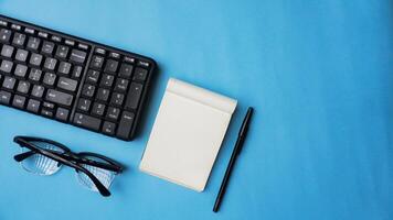 Arbeitsplatz Schreibtisch mit Notizbücher, Stifte, Klaviatur, und Brille auf Blau Hintergrund foto