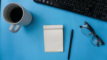 Arbeitsplatz Schreibtisch mit Notizbücher, Klaviatur, Stifte, Gläser, und Kaffee auf Blau Hintergrund foto