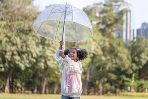 heiter Kind Mädchen mit ein Regenschirm spielen mit Regen im das Park, süß wenig Kind Mädchen spielen draußen im das Garten foto
