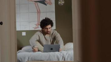 Der junge Mann aus dem Nahen Osten sitzt auf dem Bett, hat einen Laptop vor sich, unterhält sich online, passt die Haare an und redet foto