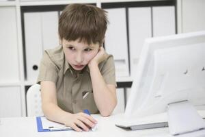 traurig Schüler tut Hausaufgaben. verärgert Junge hinter Schreibtisch mit Stift und Notizbuch.das wenig Junge denkt Über das Aufgabe foto