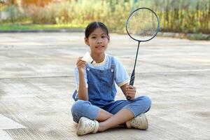 asiatisch Mädchen Sitzung mit gekreuzten Beinen auf das Zement Fußboden Hand halten ein Weiß Federball. das andere Hand hält ein Badminton Schläger. sie ausgeruht während spielen Badminton draußen auf ihr Tag aus. foto