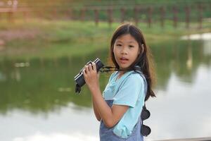asiatisch Mädchen Verwendet Fernglas zu beobachten das Natur, Vögel, und Tiere von das National Park während Camping mit ihr Familie. mit das Konzept von Lernen draußen das Klassenzimmer Natur lernen, Erkundung. foto