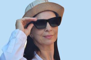 Porträt von ein kaukasisch Brünette Frau. ziemlich Frau tragen ein Hut, Weiß Hemd und Sonnenbrille auf ein Blau Hintergrund. foto