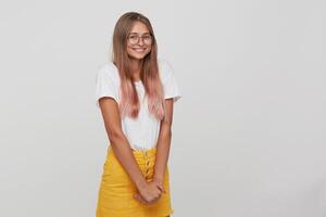 Nahansicht von glücklich schüchtern jung Frau mit lange gefärbt Pastell- Rosa Haar trägt t Shirt, Gelb Rock und Brille Stehen, lächelnd und achselzuckend ihr Schultern isoliert Über Weiß Hintergrund foto