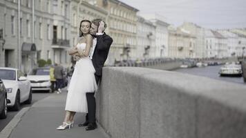 schön Jungvermählten. Aktion. ein Paar von Liebhaber, das Braut im ein fest Weiß Kleid und das Bräutigam im ein passen mit lange Haar Pose auf das Straße Nächster zu das Damm und schön historisch Gebäude im foto