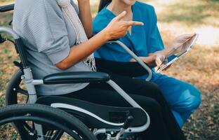 Alten asiatisch Senior Frau auf Rollstuhl mit asiatisch Vorsichtig Pflegekraft und ermutigen geduldig, Gehen im Garten. mit Pflege von ein Pflegekraft und Senior Gesundheit Versicherung im Sonne Licht foto