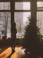 ai generiert schön jung Frau in der Nähe von das Fenster mit Weihnachten Baum beim Zuhause foto