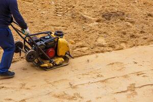 Arbeiter mit vibrierend Teller Verdichter zum Verdichtung Sand während Pfad Konstruktion. foto