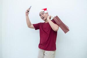 jung asiatisch Mann tragen ein Santa claus Hut halten ein Smartphone und ein Einkaufen Tasche mit Ausdrücke von lächeln, Schock, und Überraschung, isoliert gegen ein Weiß Hintergrund zum visuell Kommunikation foto