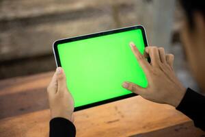 spotten oben Foto von ein schließen oben Schuss mit ein mans Hand halten ein iPad Tablette mit ein Grün Bildschirm gegen das Hintergrund von ein Holz Cafe Tabelle