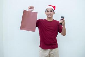 jung asiatisch Mann tragen ein Santa claus Hut halten ein Smartphone und ein Einkaufen Tasche mit Ausdrücke von lächeln, Schock, und Überraschung, isoliert gegen ein Weiß Hintergrund zum visuell Kommunikation foto