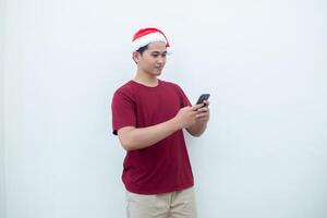 jung asiatisch Mann tragen ein Santa claus Hut halten ein Smartphone und ausdrücken ein lächeln, Schock und Überraschung isoliert durch ein Weiß Hintergrund zum visuell Kommunikation foto