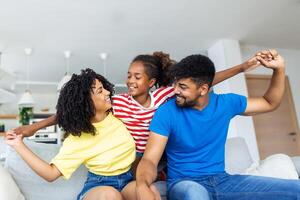 heiter afrikanisch Mutter und Vater spielen mit Tochter beim heim. süß Mädchen genießen Sitzung auf Vater Schulter während suchen beim Kamera. jung Familie haben Spaß zusammen auf das Sofa beim heim. foto