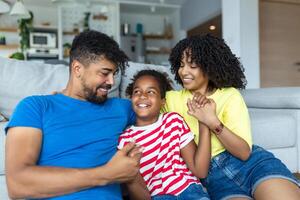 multiethnisch Familie Sitzung auf Couch im Leben Zimmer beim Zuhause genießen kostenlos Zeit zusammen auf Wochenende kommunizieren mit jeder andere. schön Tochter Umarmung Mutter hat Spaß mit Eltern drinnen foto