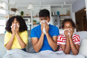 Porträt von krank jung schwarz Familie von drei Menschen weht flüssig Nasen während Sitzung zusammen auf das Sofa mit Servietten und bedeckt mit Decke. foto