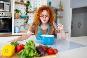 heiter jung ziemlich weiblich im Schürze bereiten Mittagessen und Geruch Gericht im Küche beim heim. Frau Kochen Abendessen zum Familie beim Neu Rezept beim heim. foto