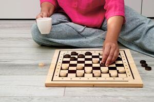 Frau macht ein Bewegung im ein Spiel von Dame beim Zuhause während Sitzung auf das Fußboden und halten ein Becher von Tee foto