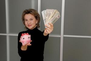 ein zufrieden Frau hält aus ein Ventilator von ihr Banknoten und hält ein Schweinchen Bank im ihr Hände foto