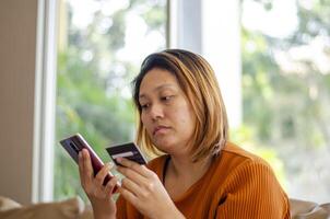 asiatisch Frau fallen Opfer zu finanziell Transaktion, Hand halten auf Anerkennung Karte und Handy, Mobiltelefon Telefon foto