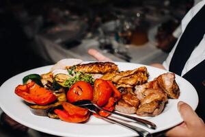 im das Abend, das Restaurant dient ein heiß Gericht von Grill mit Gemüse.gebraten Fleisch mit Tomaten und andere Gemüse auf ein Teller im ein Restaurant. foto