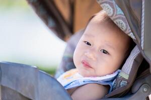 Baby entspannend im ein Kinderwagen außen. Kleinkind Junge mit leicht Lächeln ruht, suchen aus mit neugierig Augen, genießen das Komfort und Sehenswürdigkeiten von ein gemächlich Tag draußen. asiatisch Kind alt 1 Jahre alt. foto