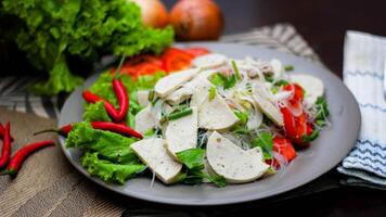 würzig Glas Nudel Salat mit Vietnamesisch Würstchen serviert im ein grau Teller stellen auf ein hölzern Tabelle und verschiedene Gemüse. foto