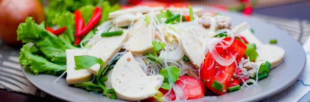 würzig Glas Nudel Salat mit Vietnamesisch Würstchen serviert im ein grau Teller stellen auf ein hölzern Tabelle und verschiedene Gemüse. foto