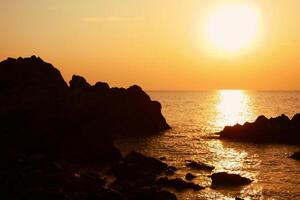 das Sonne ist Rahmen auf das Meer mit Felsen im das Vordergrund, schwarz Silhouetten auf das Felsen, Licht reflektieren aus das Meer. foto