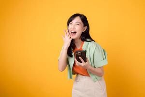 Schönheit aufgeregt asiatisch jung 30er Jahre Frau Ausdruck schreien bekannt geben Geste während halten Smartphone isoliert auf Gelb Hintergrund. attraktiv süß weiblich erstaunt sprechen Jubel Rabatt Werbung. foto