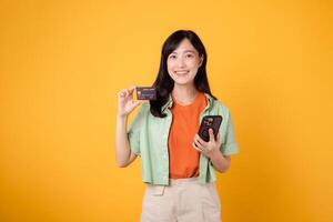 Nervenkitzel von Einkaufen von Handy, Mobiltelefon mit ein fesselnd jung asiatisch Frau im ihr 30er Jahre, elegant gekleidet im Orange Hemd und Grün Jumper, mit Smartphone zu Show Anerkennung Karte auf Gelb Studio Hintergrund. foto