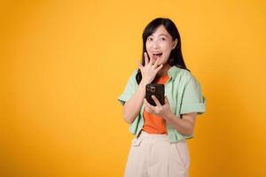 erkunden das Charme von Überraschung mit ein heiter jung asiatisch Frau im ihr 30er Jahre, gekleidet im ein Orange Shirt, mit ihr Smartphone, Mund geschlossen, auf ein Gelb Studio Hintergrund. App Smartphone Konzept. foto