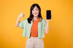 Nervenkitzel von ein Neu Handy, Mobiltelefon Anwendung mit ein jung asiatisch Frau im ihr 30er Jahre, gekleidet im Orange Hemd und Grün Jumper, präsentieren Smartphone Bildschirm mit ein Daumen hoch Geste auf Gelb Studio Hintergrund. foto