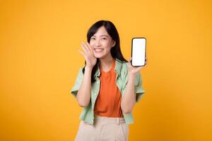jung asiatisch Frau im ihr 30er Jahre, gekleidet im Orange Hemd und Grün Jumper, Anzeigen Smartphone Bildschirm mit ein schreien Mund auf Gelb Studio Hintergrund. Neu Handy, Mobiltelefon Gerät und online Einkaufen Konzept. foto