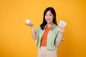 glücklich jung asiatisch Frau im ihr 30er Jahre, tragen Orange Hemd und Grün Jumper, Vitrinen Dollar Währung und Schweinchen Bank auf Gelb Hintergrund. finanziell Geld Konzept. foto