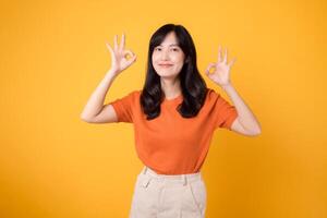Umarmung positiv Stimmung wie ein jung asiatisch Frau im ihr 30er Jahre, anziehen ein Orange Shirt, Vitrinen das okay Zeichen Geste auf ein sonnig Gelb Hintergrund. Hände Geste Konzept foto