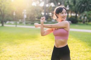 Wellness und gesund Lebensstil Porträt von 30er Jahre asiatisch Frau im Rosa Sportbekleidung. bereiten und strecken Körper und Arm Muskeln Vor Sonnenuntergang Lauf im das Park. Fitness draußen und Leben ein ausgewogen Leben. foto