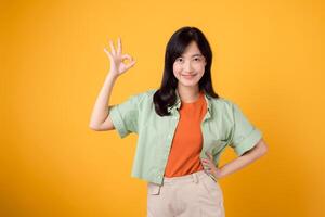 jung asiatisch Frau 30er Jahre, elegant gekleidet im Orange Hemd und Grün Jumper. ihr liebenswert okay Hand Geste und sanft lächeln, einstellen gegen ein Gelb Hintergrund, verraten das Schönheit von Körper Sprache. foto
