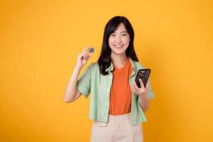 jung asiatisch Frau 30er Jahre, elegant gekleidet im Orange Hemd und Grün Jumper, zeigen Krypto Währung Münze während halten Smartphone auf Gelb Hintergrund. Zukunft Finanzen Konzept. foto