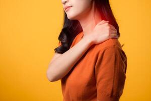 unterstützend asiatisch Frau im ihr 30er Jahre, tragen ein Orange Shirt, hält ihr Schmerzen Schulter auf Gelb Hintergrund. Hals schmerzen Therapie medizinisch Büro Syndrom Konzept. foto