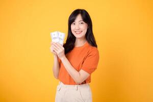 heiter jung asiatisch Frau im ihr 30er Jahre, halten Kasse Geld Dollar, Stehen auf beschwingt Gelb Hintergrund. foto