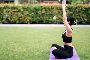 Porträt passen jung asiatisch Frau 30er Jahre tragen schwarz Sportbekleidung Hör mal zu entspannend Musik- während tun Yoga Übung auf Yoga Matte im Grün Natur Park. Wohlbefinden Konzept. foto