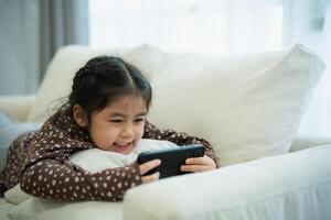 asiatisch Kind Mädchen suchen mit und berühren Handy, Mobiltelefon Telefon Bildschirm auf Couch Sofa. Baby lächelnd komisch Zeit zu verwenden Handy, Mobiltelefon Telefon. auch viel Bildschirm Zeit. süß Mädchen Aufpassen Videos während Fernseher, Internet Sucht. foto