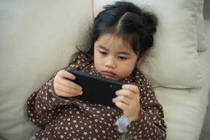 asiatisch Kind Mädchen ernst oder konzentrieren und mit Handy, Mobiltelefon Telefon Bildschirm auf Couch Sofa. Baby lächelnd komisch Zeit verwenden Handy, Mobiltelefon Telefon. auch viel Bildschirm Zeit. Mädchen Aufpassen Videos während Fernseher, Internet Sucht. foto