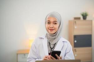 Muslim Islam asiatisch Arzt Mann tragen Hijab und Stethoskop Arbeiten mit Laptop Computer und mit Handy, Mobiltelefon Telefon, Smartphone. weiblich Arzt Arbeit beim Zuhause Büro Krankenhaus. Gesundheit Krankenhaus Konzept. foto