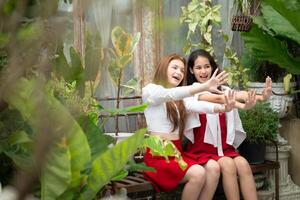 zwei jung asiatisch Frauen im rot und Weiß Kleider Sitzung auf ein hölzern Bank im das Garten. foto